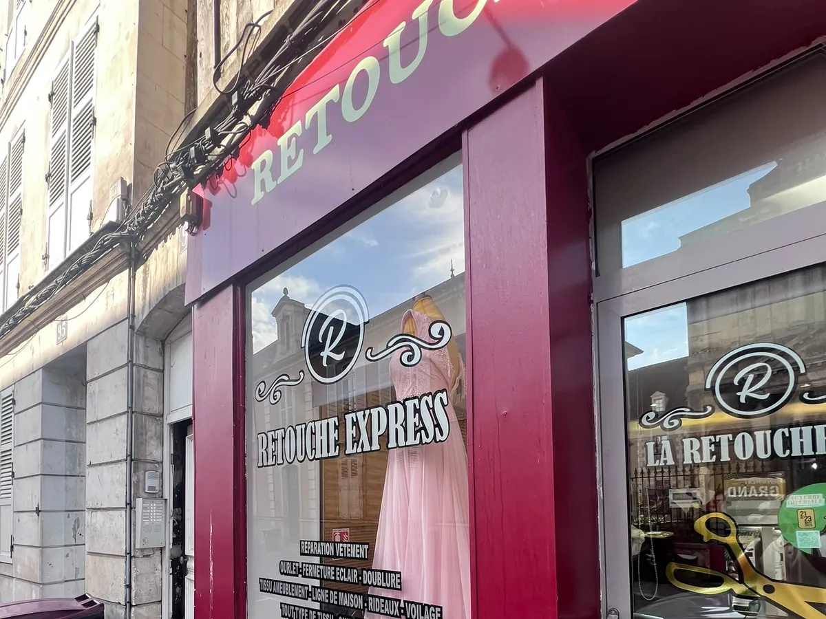 Devanture de Retouche Express vue de côté, une robe rose en vitrine et les horaires sur la porte